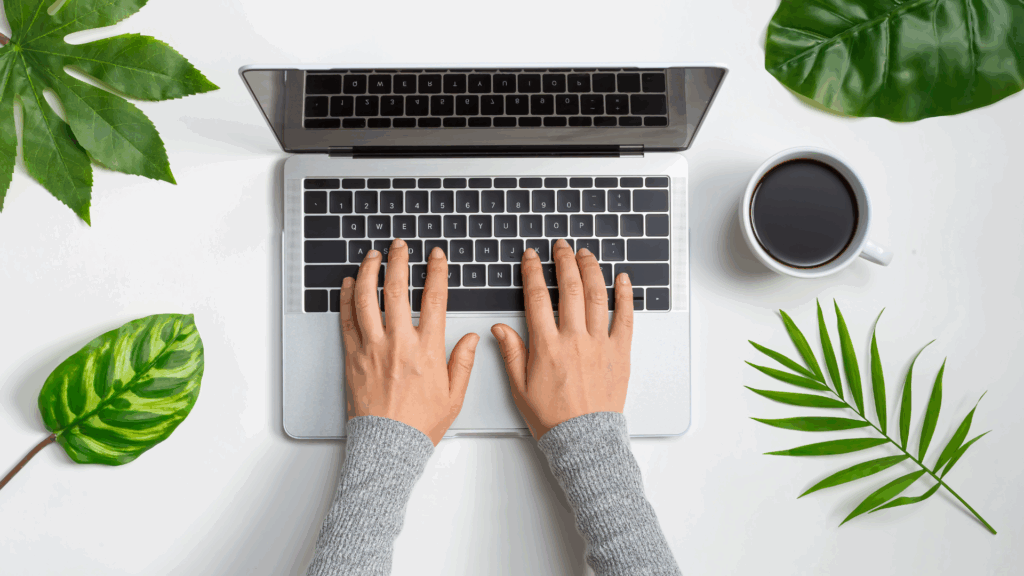 A top-down view of a close-up of hands typing on a laptop on a desk with a cup of coffee.